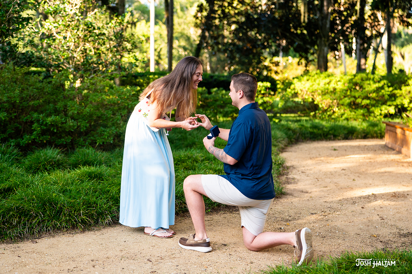 A man kneeling on one knee proposes to a woman in a blue dress, smiling as she accepts the engagement ring in a lush garden setting.