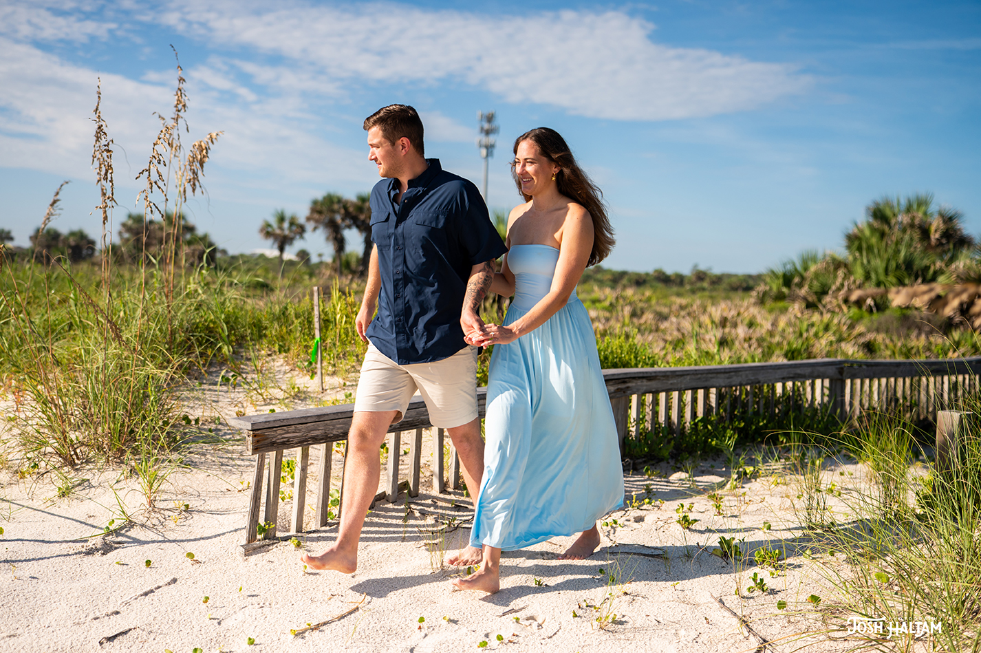 A couple walking hand in hand on a sandy beach, surrounded by tall grass and palm trees, with a clear blue sky above.