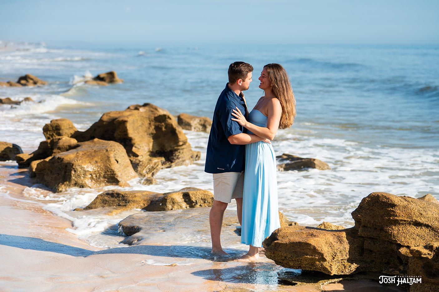 A couple embraces each other on a sandy beach with rocks and ocean waves in the background, surrounded by a sunny sky.