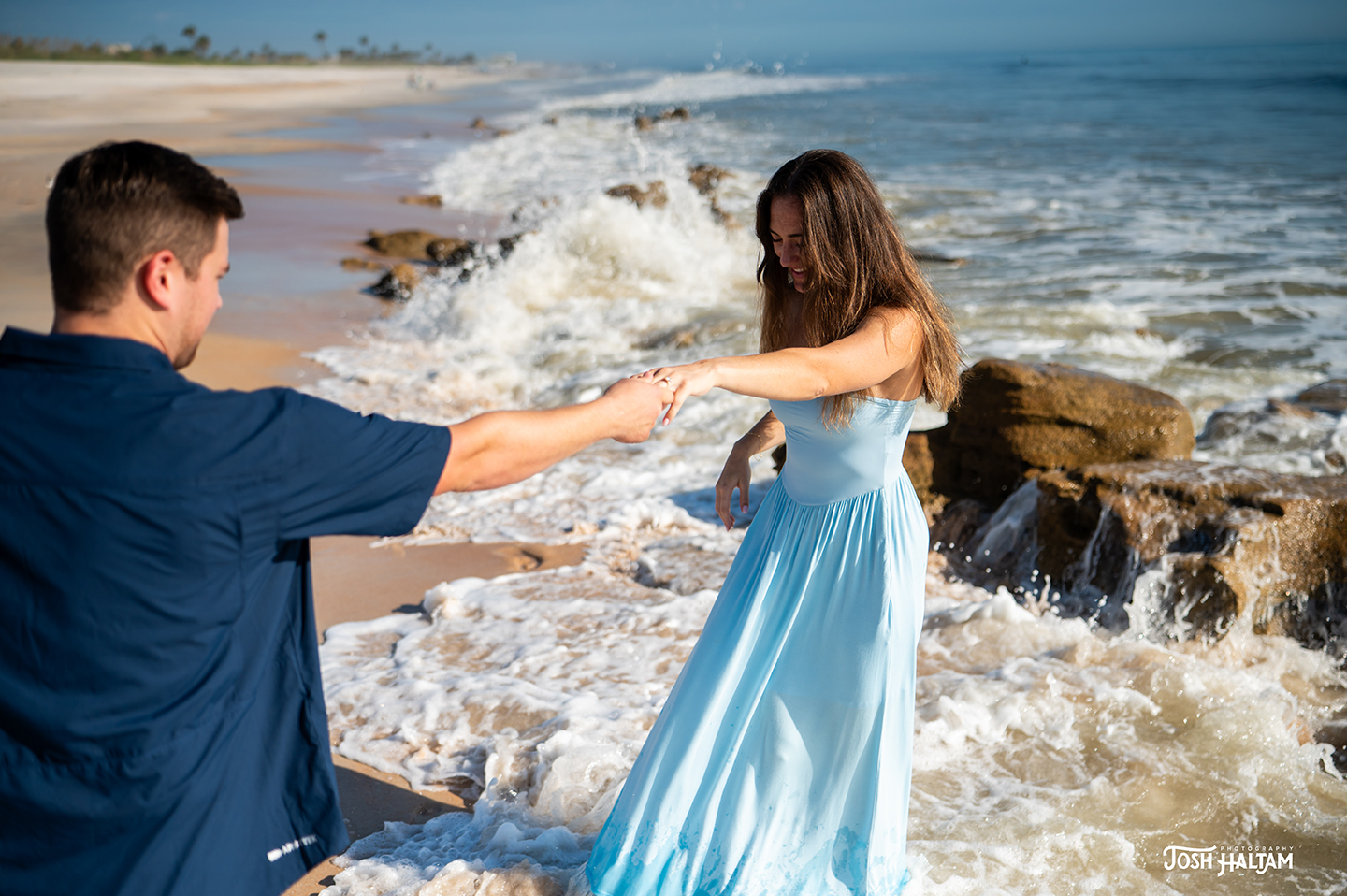 A man and woman holding hands on a beach, with the woman in a light blue dress playing in the waves near rocky shore.