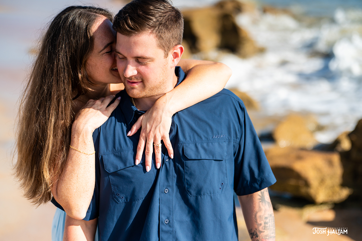A couple enjoying a romantic moment on the beach, with the woman playfully kissing the man on the cheek, surrounded by a scenic coastal background.