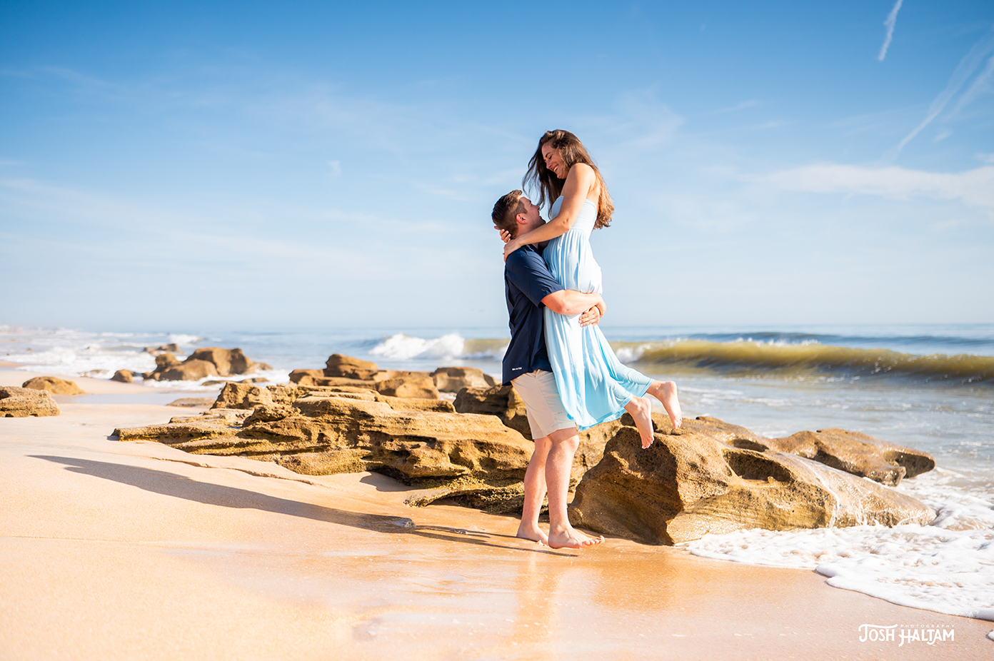 A couple embracing on a beach, with the woman in a blue dress being lifted by the man. In the background, gentle waves and rocky formations are visible under a clear blue sky.