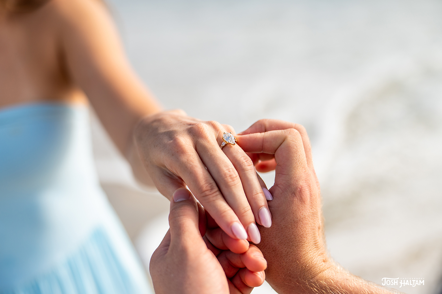 A close-up image of a hand placing an engagement ring on another hand, with a beach in the background.
