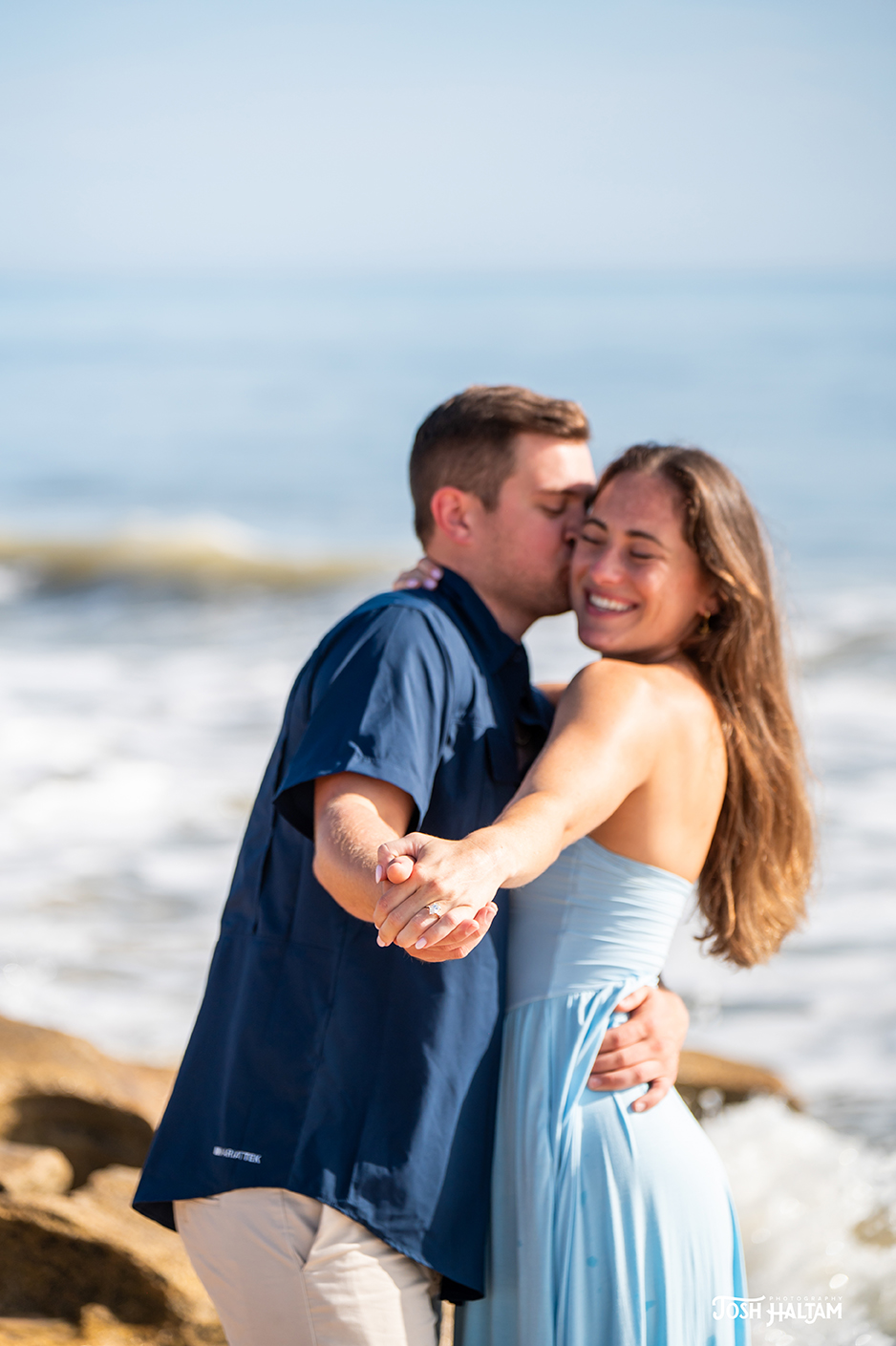 A couple shares a joyful moment on the beach, with the man kissing the woman's cheek as she smiles, holding hands in front of the ocean.