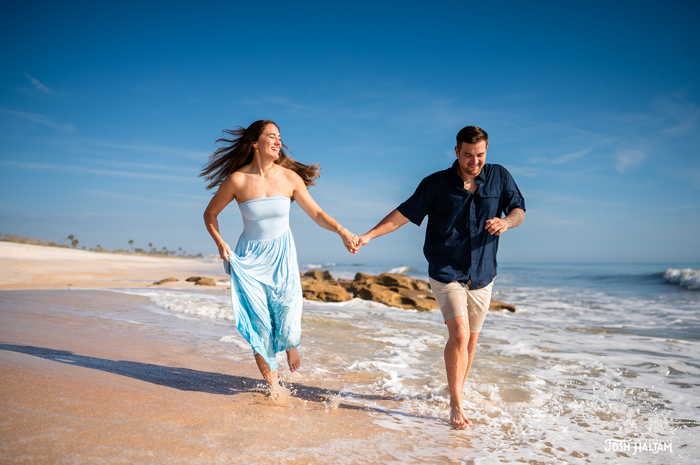A couple joyfully running along a beach, holding hands and splashing through the waves. The woman is wearing a light blue dress, and the man is in a blue shirt and beige shorts, set against a clear blue sky.