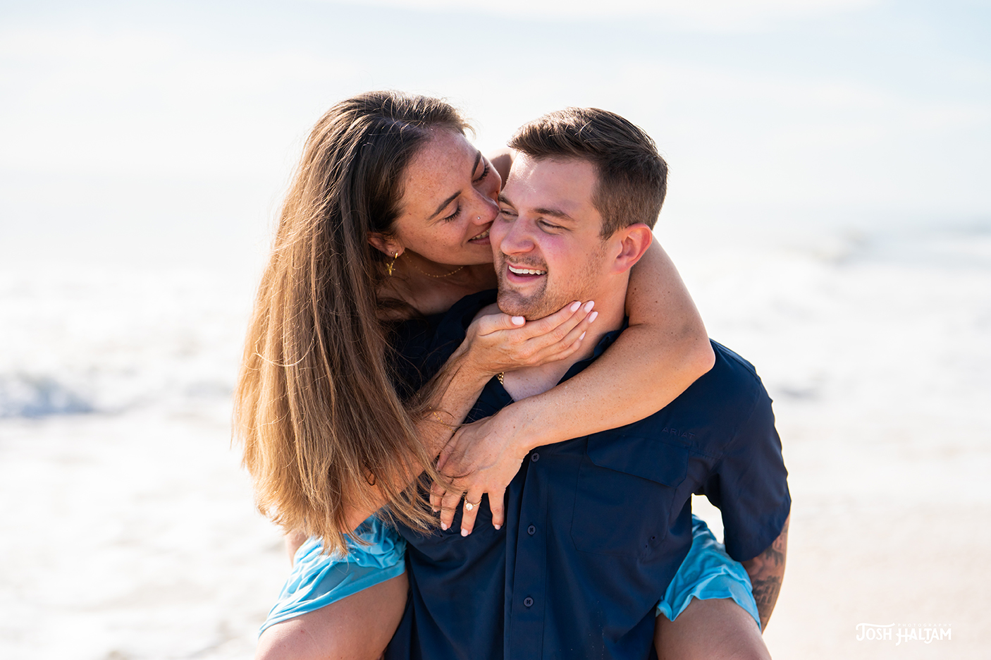A couple playfully embracing at the beach, with the woman sitting on the man's shoulders and smiling while the man laughs.