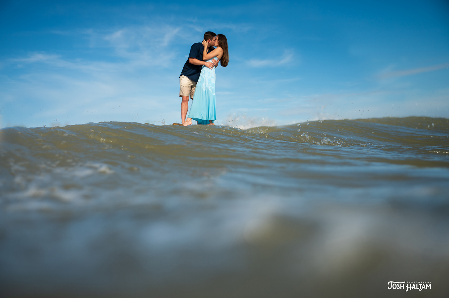 A couple embracing and kissing on a shallow wave at the beach, under a clear blue sky.