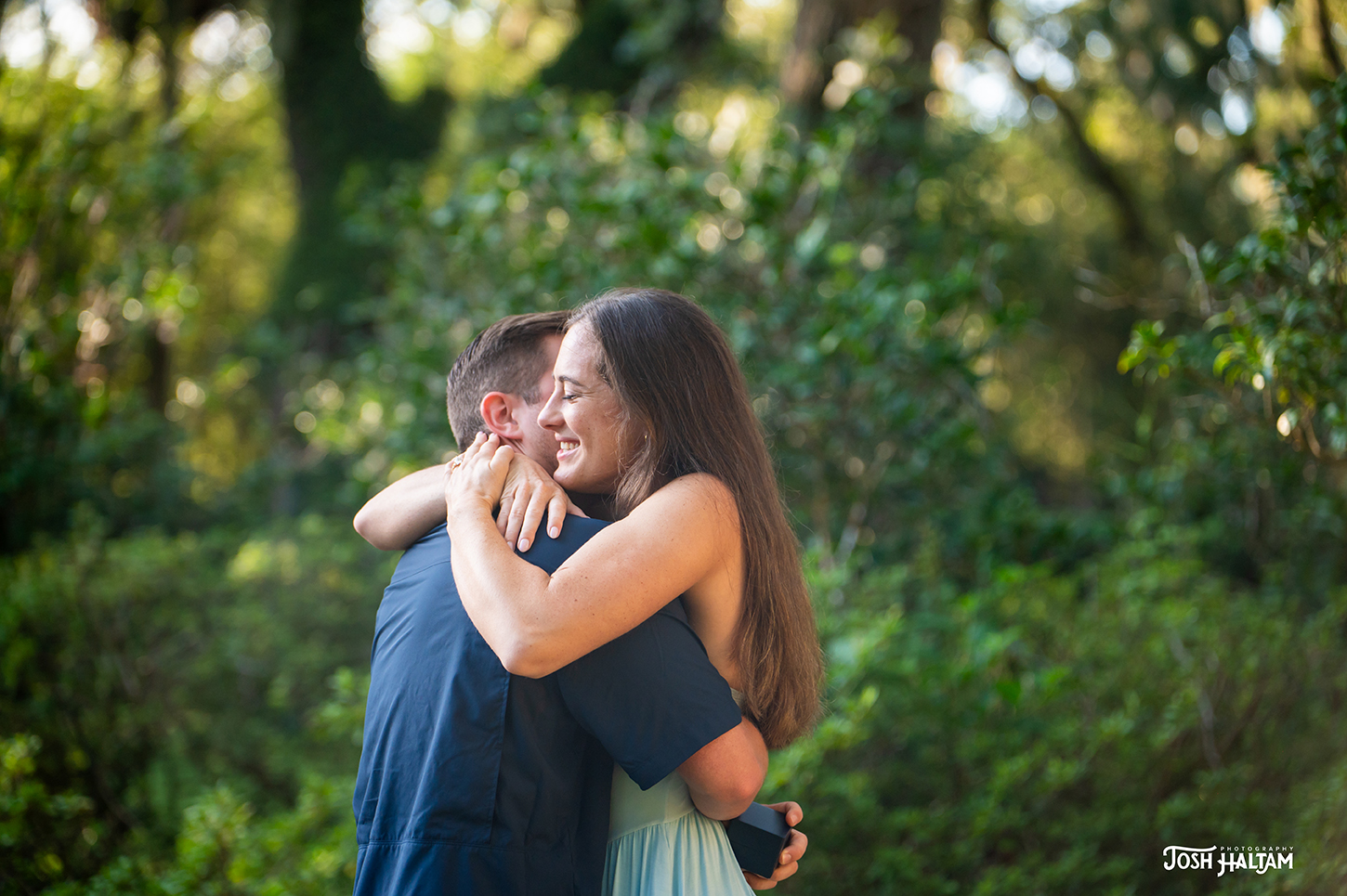 A couple embracing joyfully in a natural outdoor setting, with greenery in the background. The woman is wearing a light blue dress and appears to be smiling as she hugs the man, who is wearing a dark shirt and holding a small box.