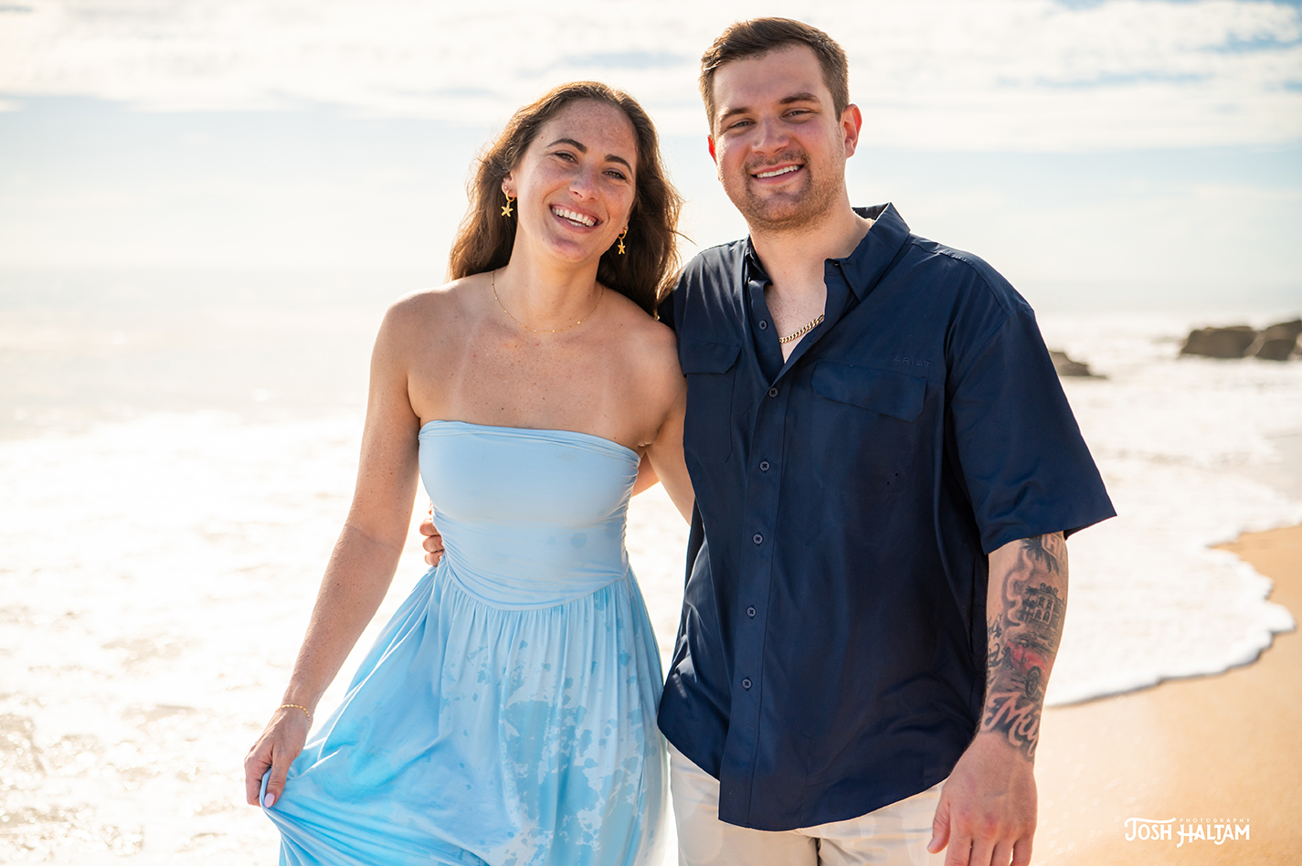 A smiling couple stands together on a beach, with the ocean waves in the background. The woman is wearing a light blue strapless dress, and the man is wearing a dark blue button-up shirt.