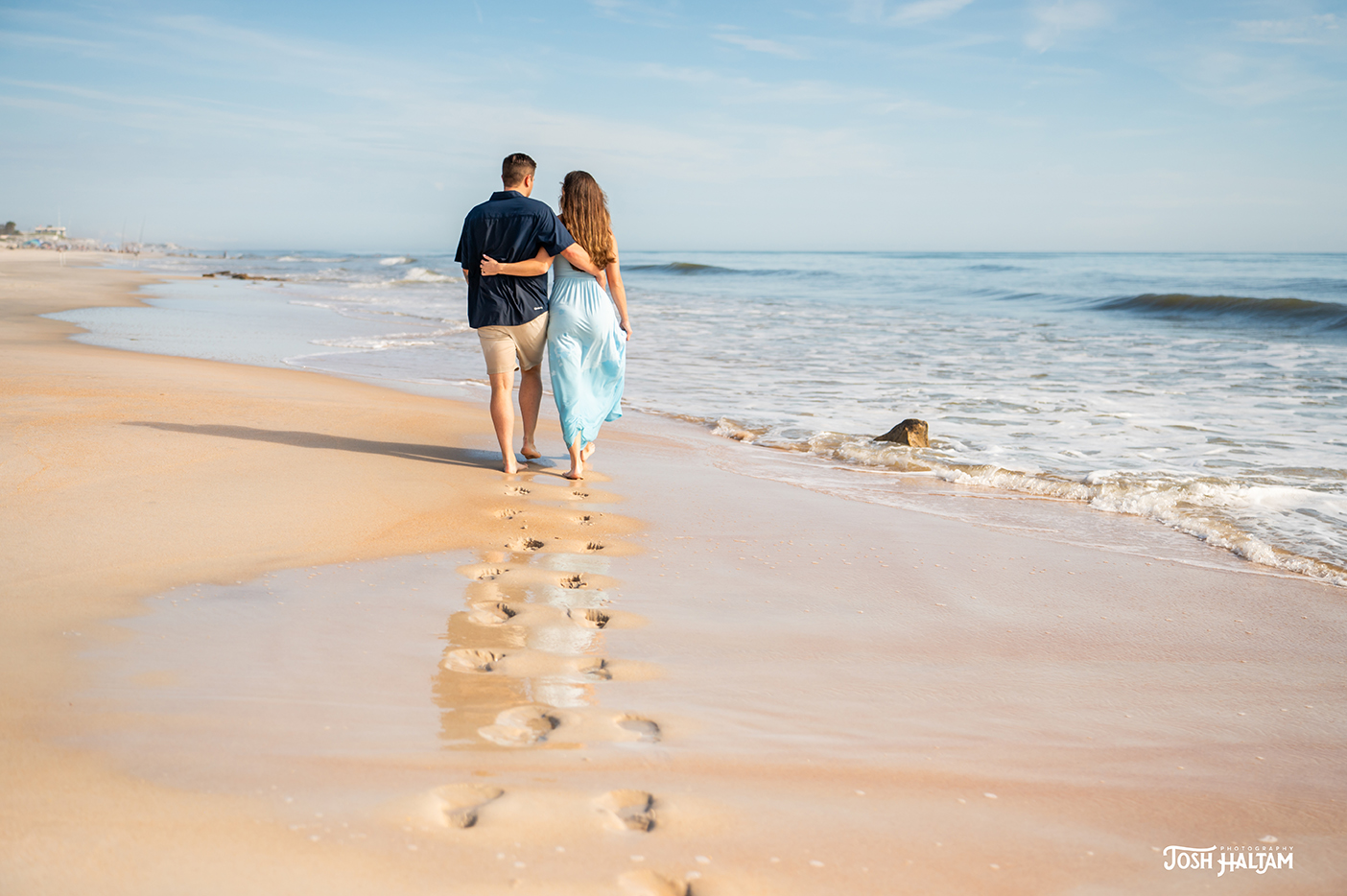 A couple walking hand in hand along a sandy beach, with gentle waves lapping at their feet.
