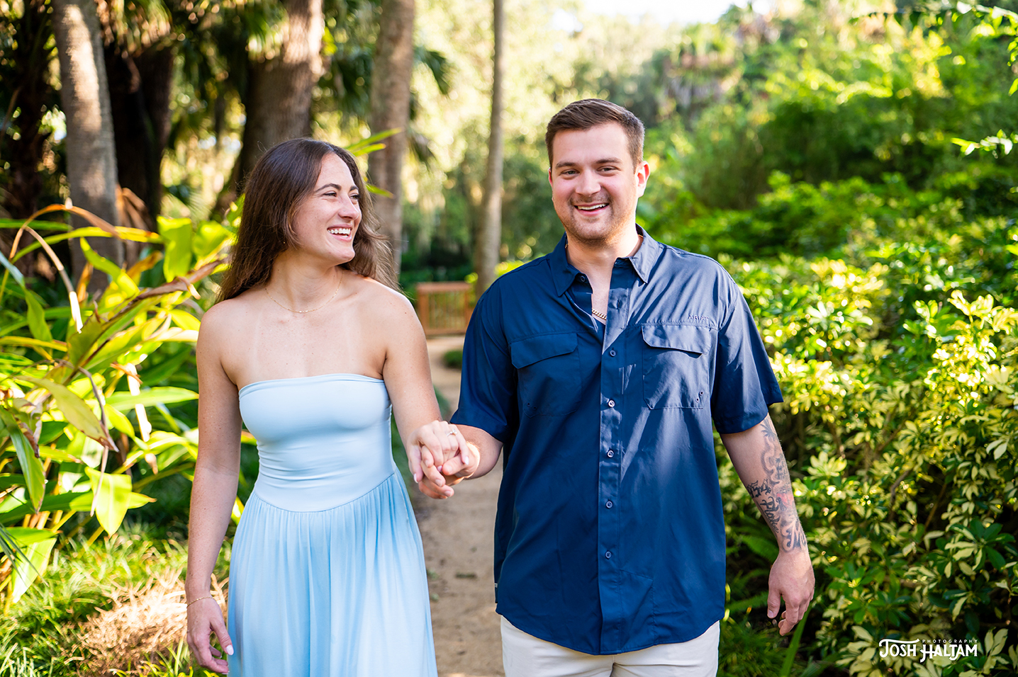 A happy couple walking hand in hand along a path surrounded by lush greenery. The woman is wearing a light blue strapless dress, and the man is dressed in a navy blue short-sleeve shirt and light shorts. Both are smiling as they enjoy their time together.