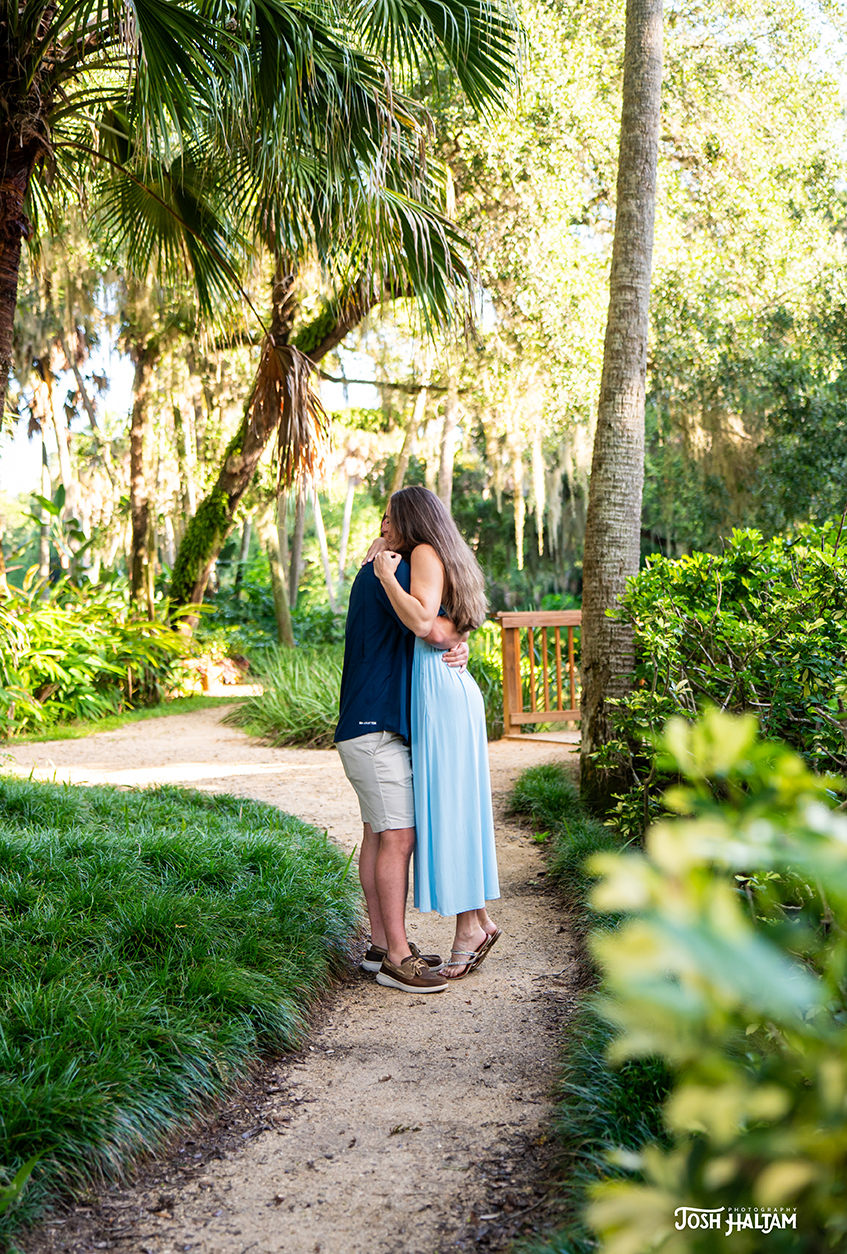 A couple embraces on a pathway surrounded by lush greenery and palm trees, with a wooden bridge visible in the background.
