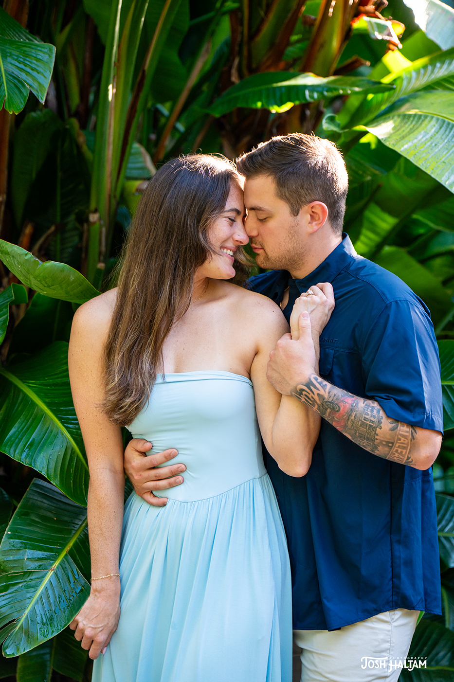 A couple embracing in a tropical setting, surrounded by lush green plants, with their foreheads touching and smiling at each other.