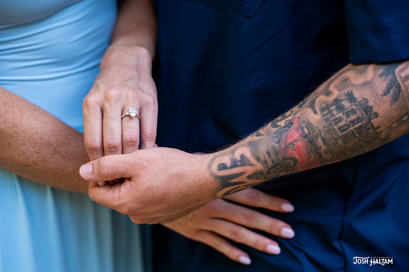 Close-up of a couple's hands holding each other, showcasing an engagement ring on the woman's finger and intricate tattoos on the man's arm.