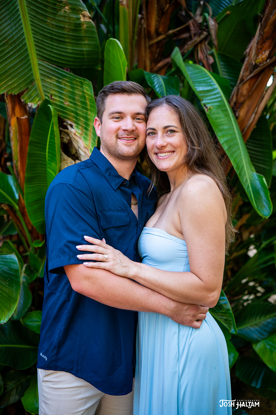 A couple embraces in front of lush tropical greenery. The man wears a navy blue shirt, and the woman is in a light blue dress, smiling at the camera.