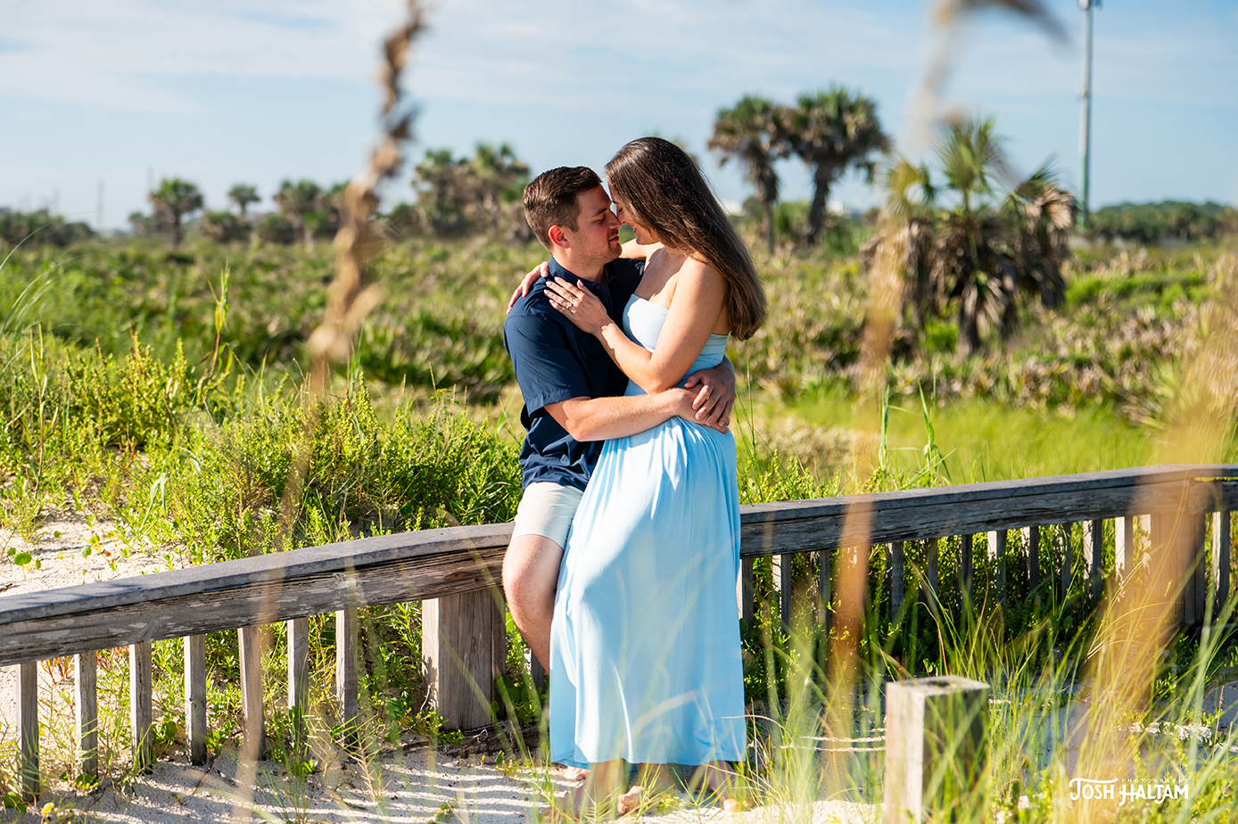 A couple sitting closely on a wooden bench in a lush green outdoor setting, with the woman in a light blue dress and the man in a navy shirt.